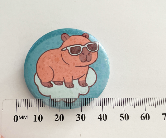 Button badge with a caybara wearing sunglasses on a blue background, measured against a ruler.