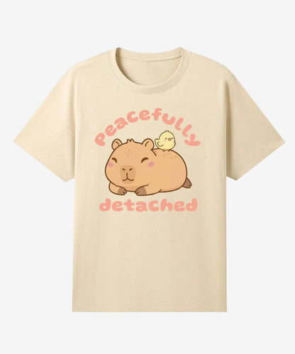 Beige t-shirt with a cartoon capybara and chick design and text 'Peacefully detached' on a white background.